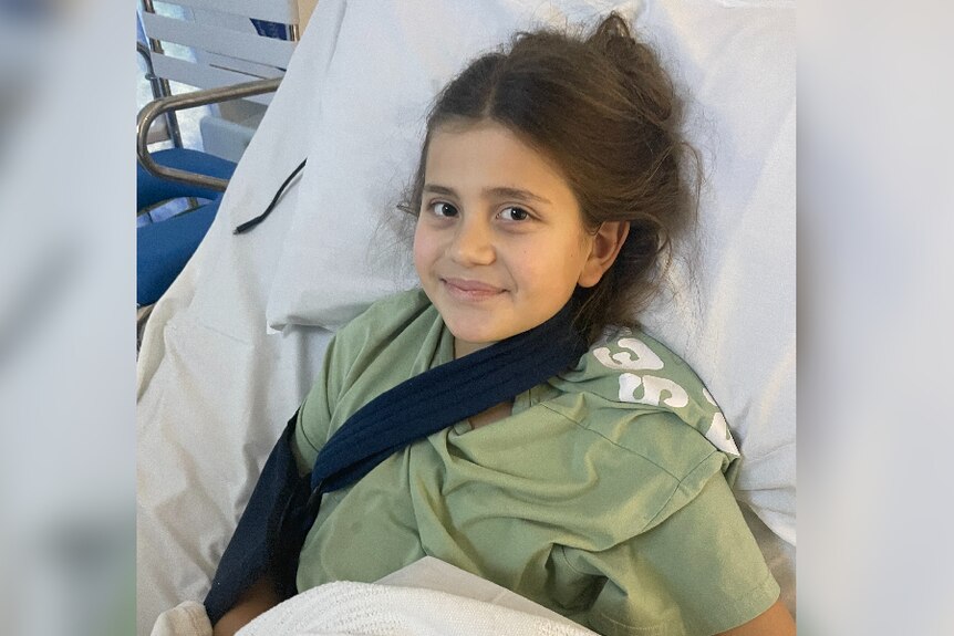 A girl in a green shirt and brace smiles while lying in a hospital bed