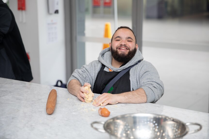 A male day program participant is smiling broadly as he kneads dough, at a kitchen bench covered with flour and egg shells.