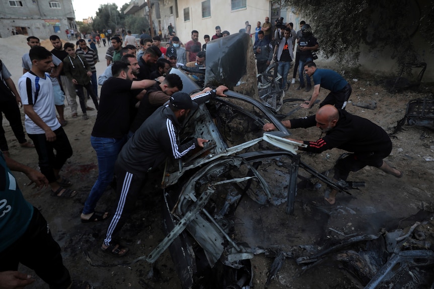 Palestinians gather around the shell of a car destroyed by an air strike.