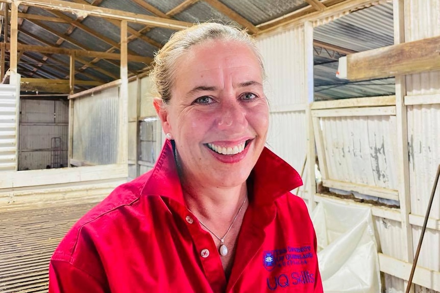 A women wearing a red collared shirt smiles at the camera while sitting in a shearing shed