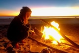 A woman crouches next to a campfire on a beach at sunset