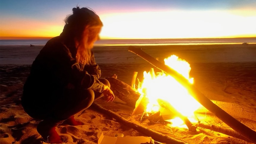 A woman crouches next to a campfire on a beach at sunset