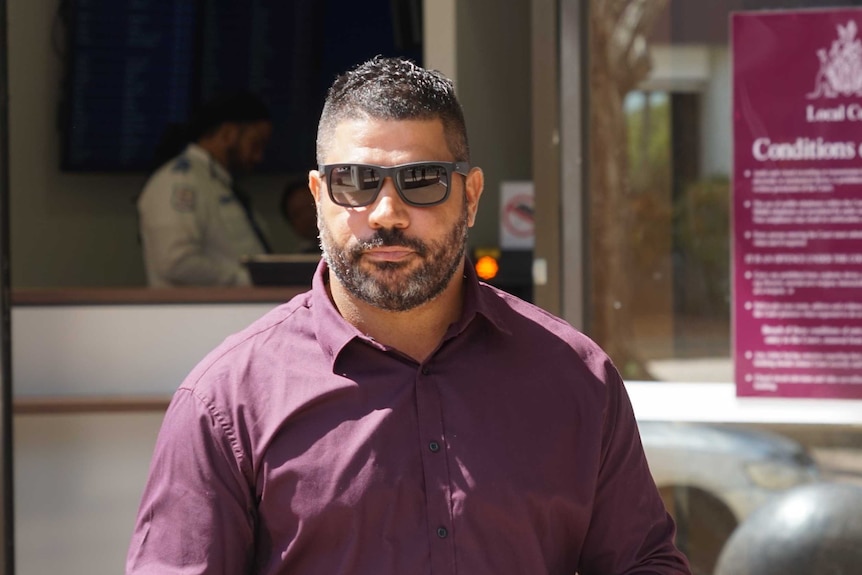 A man with dark glasses on walks across a streetscape.