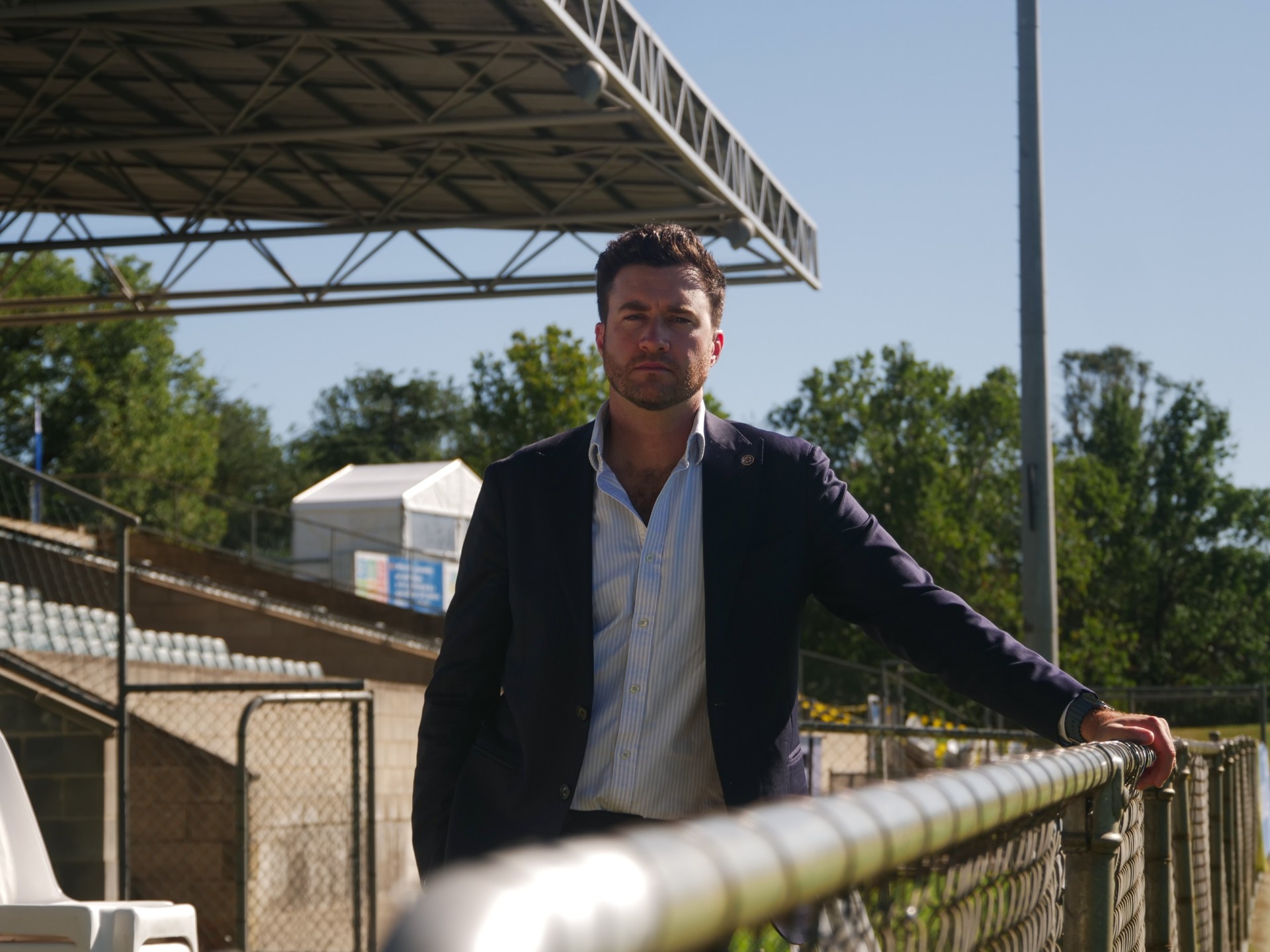 Ben in a suit standing in front of a grandstand, looking at the camera and leaning with one arms against a fence.