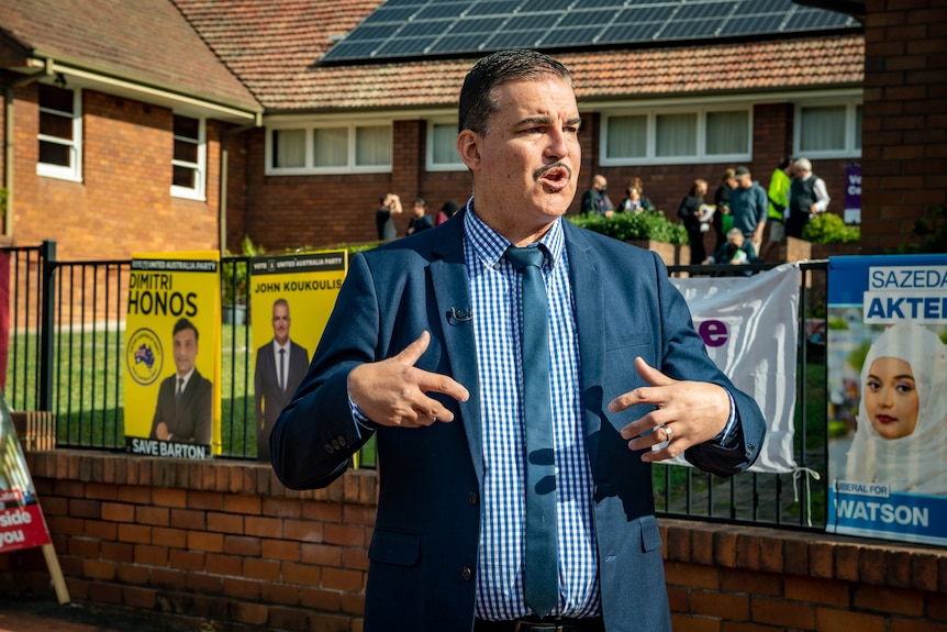 John Koukoulis speaking in front of a fence that is decorated with campaign posters from the UAP and Liberals.