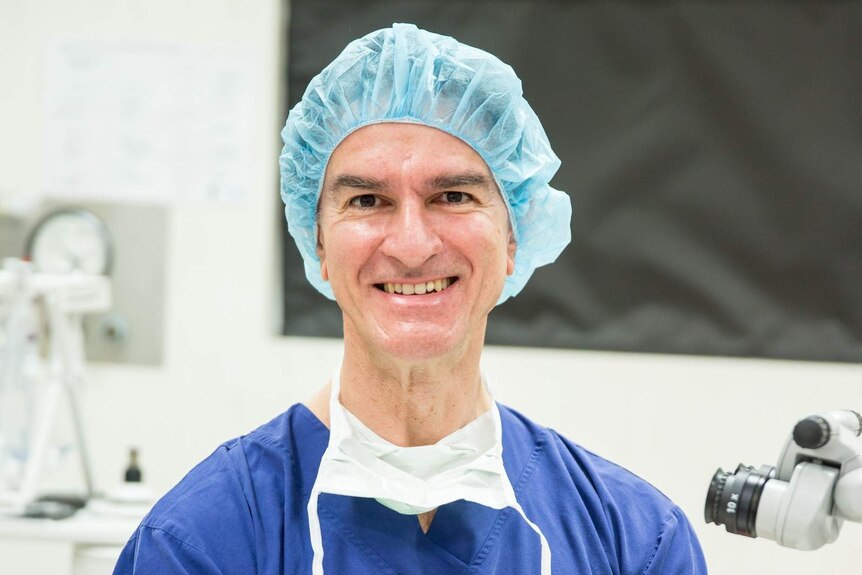 A surgeon wearing a protective cap and a blue gown stands in an operating theatre.