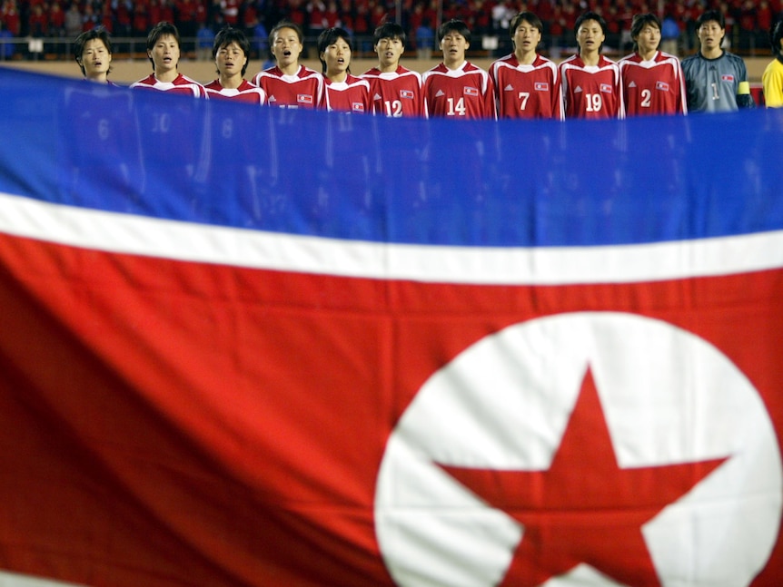 A soccer team wearing red and white stands behind a flag of red, white and blue in a line