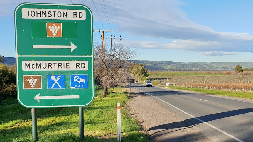 A green road sign with white text next to a country road leading through vineyards
