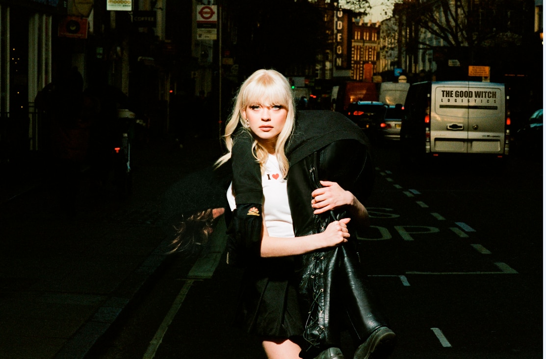 Maisie Peters stands on a dark street in a white tee shirt with someone wearing black leather thrown over her shoulder.