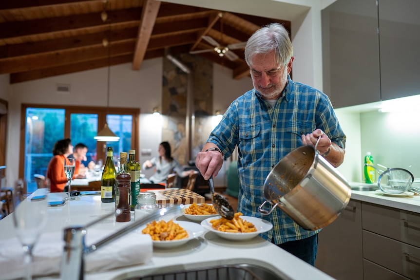 A man spoons out pasta into bowls in a kitchen, behind him people sit around a dining table.