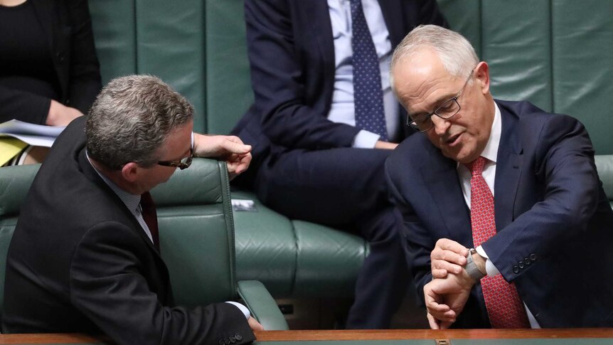 Malcolm Turnbull, wearing a red tie, leans over towards Christopher Pyne while adjusting his wrist watch.
