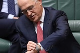 Malcolm Turnbull, wearing a red tie, leans over towards Christopher Pyne while adjusting his wrist watch.