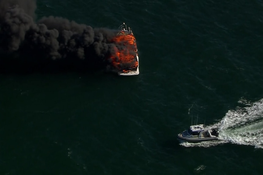 Two boats on the water, with one up in flames with black smoke billowing out.