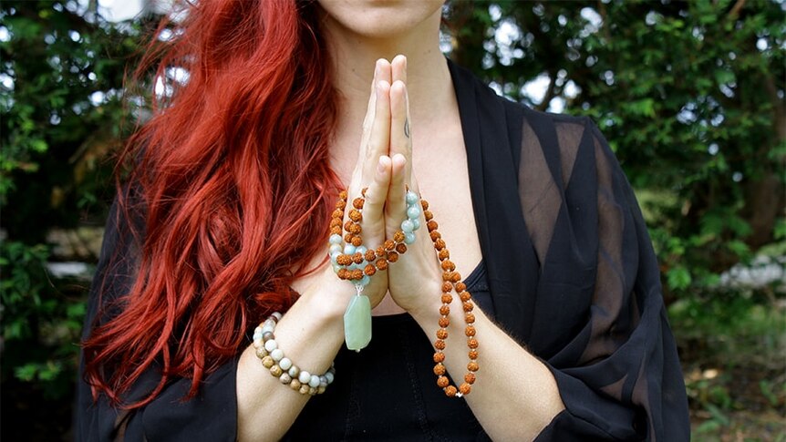 A woman with long red hair clasps her hands in prayer with beads wrapped around them