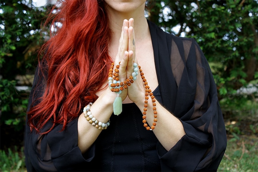 A woman with long red hair clasps her hands in prayer with beads wrapped around them