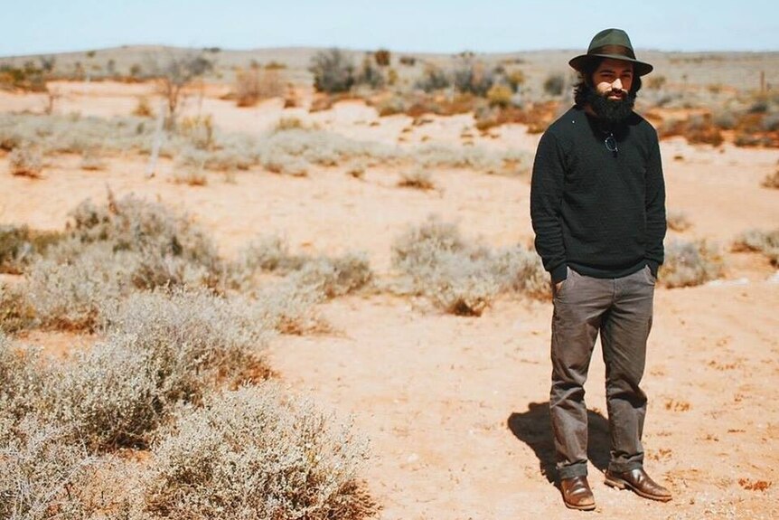 Criminal lawyer Jalal Razi, standing in an arid desert-like setting, wearing dark clothes and a brimmed hat.