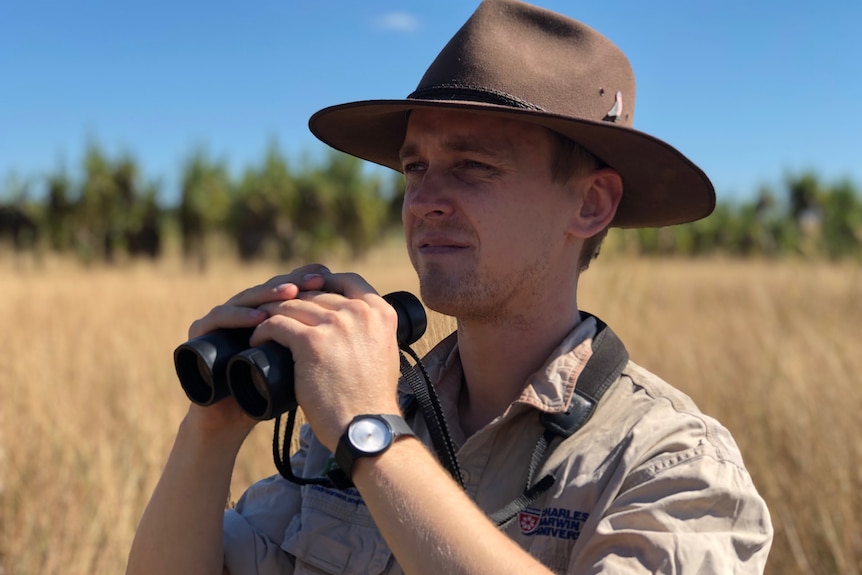 A man wearing a hat holds binoculars below his view and looks into the distance. 