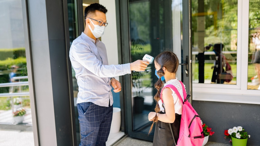 A teacher wearing the mask takes the temperature of a young girl.