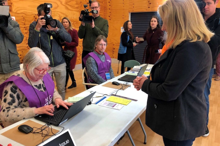 A woman standing at a table in a polling booth surrounded by media cameras.