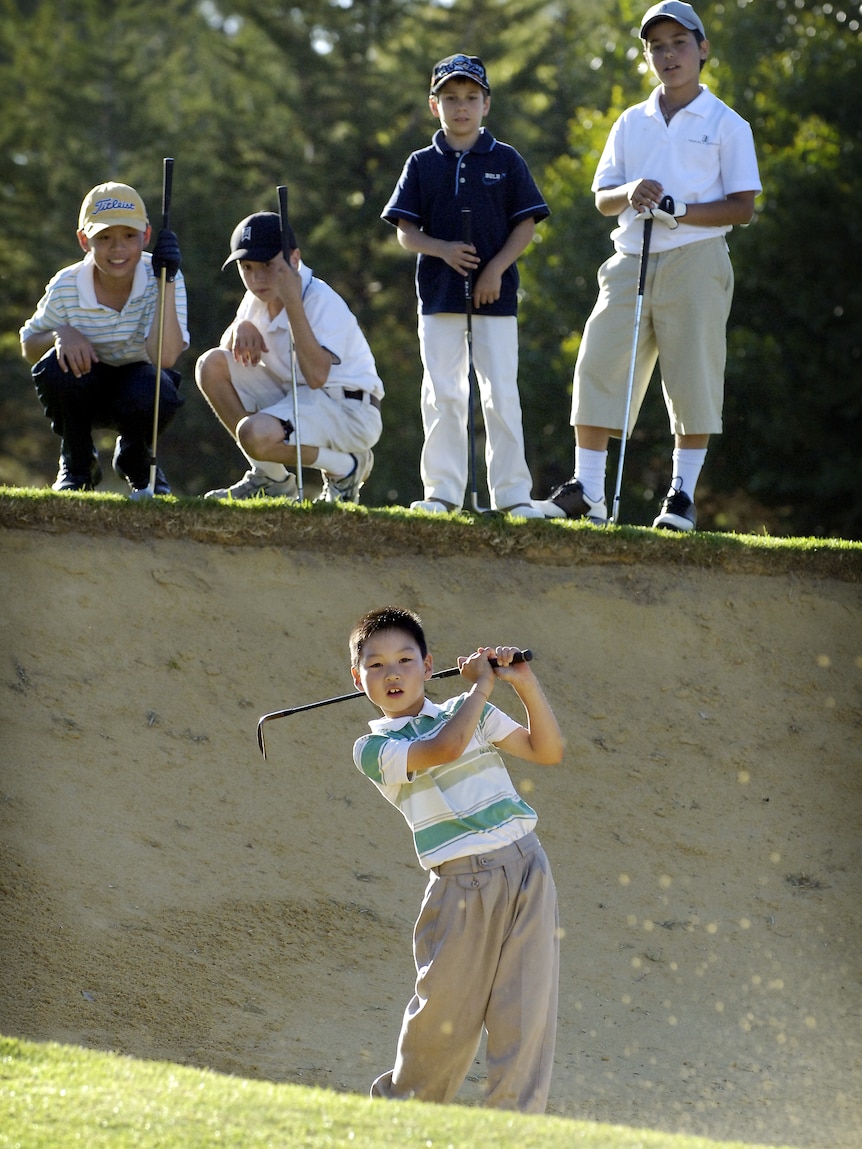 A boy wearing a polo shirt and khaki pants puts a ball out of the sand on a golf course. Four other boys watch on.