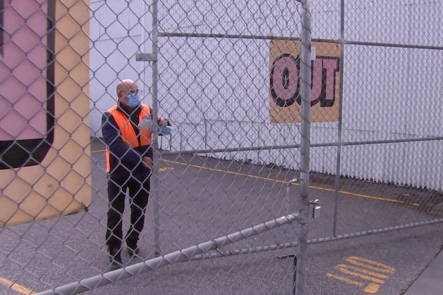 A man wearing a mask closes a gate with a sign saying OUT on it