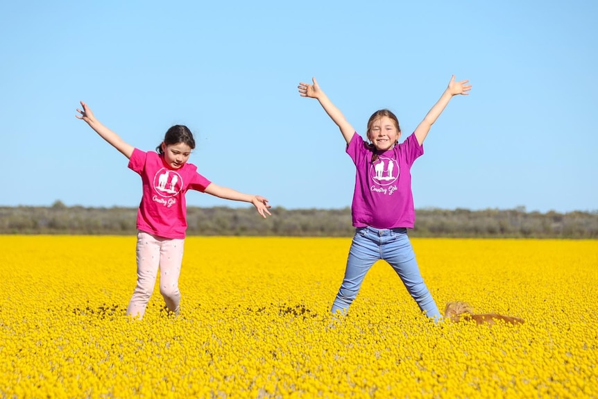 Two children wearing bright pink shirts star jump surrounded by bright yellow flowers.