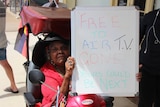 A local resident sitting in her mobility scooter holds a sign that reads, 'Free to air TV gone, yours could be next
