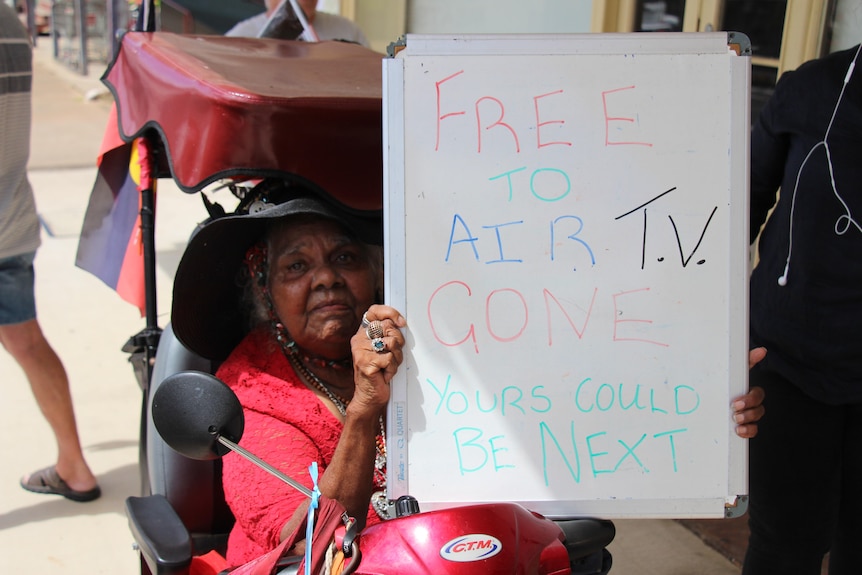 A local resident sitting in her mobility scooter holds a sign that reads, 'Free to air TV gone, yours could be next