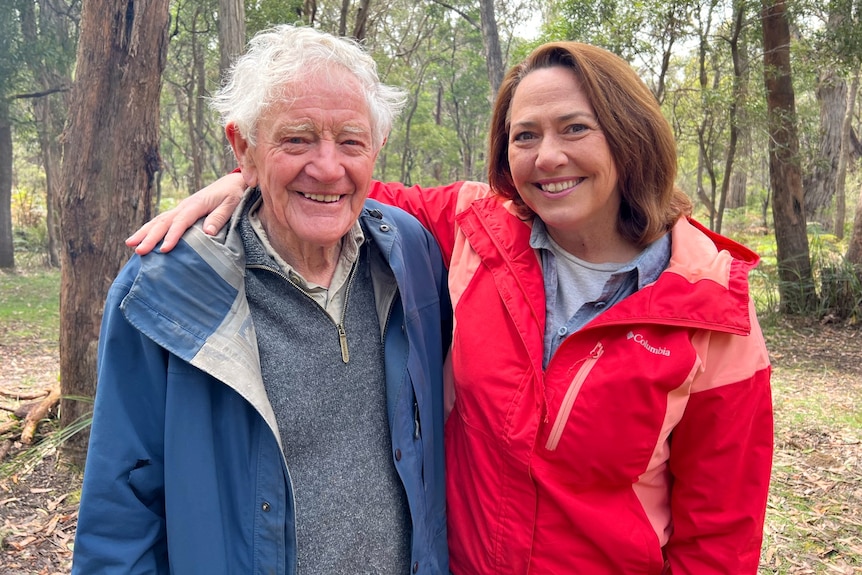 A woman in a bright pink jacket stands in a forest with her arm around an elderly man with white hair, smiling.