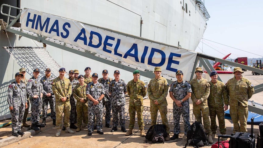 A group of Australian Defence Force troops stand in a row with a HMAS Adelaide sign in the back