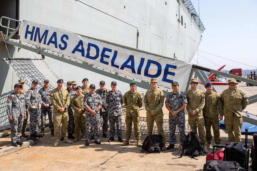 A group of Australian Defence Force troops stand in a row with a HMAS Adelaide sign in the back