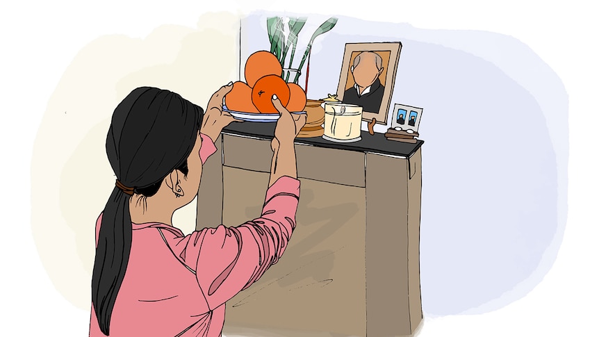 A woman with dark hair and pink shirt places a bowl of oranges on a small altar featuring a framed photo and incense