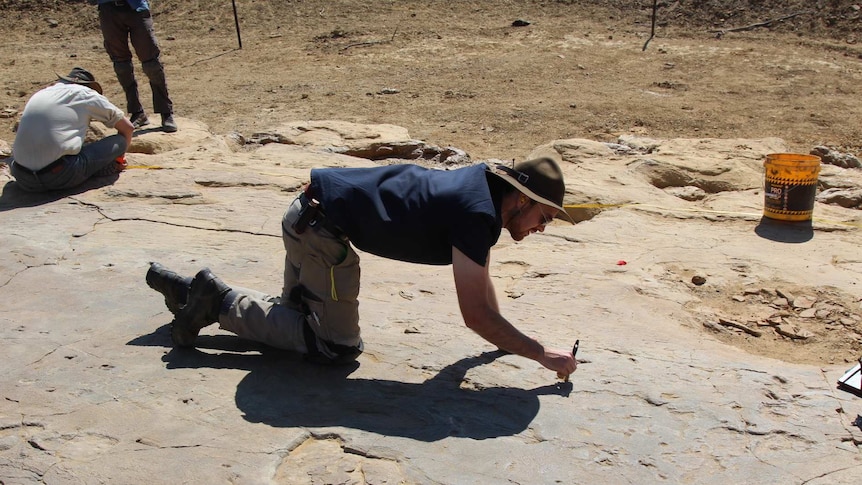 A man wearing a broad-brimmed hat crouches on pockmarked rock and uses an implement to mark the rock.