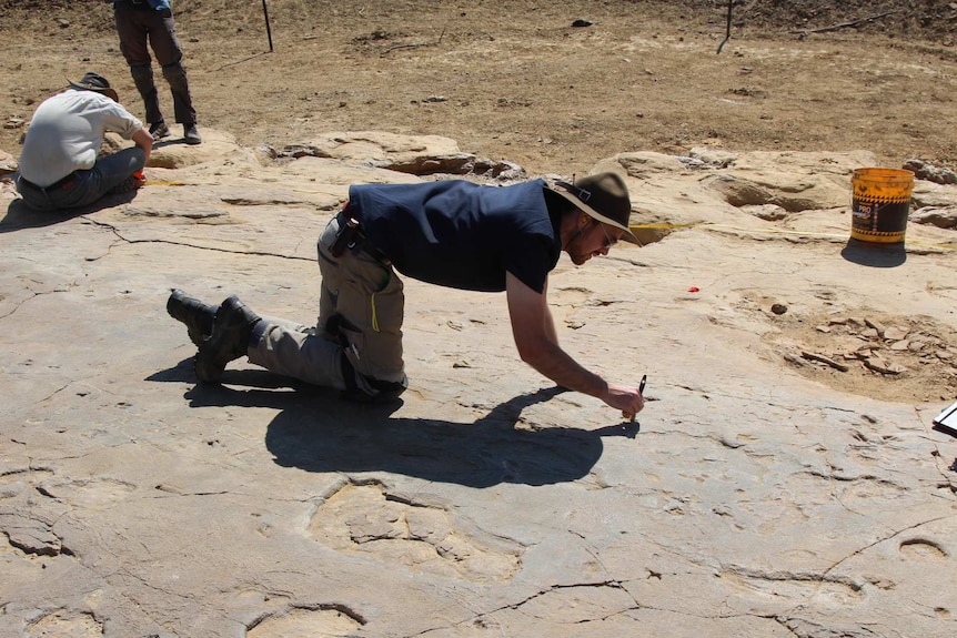 A man wearing a broad-brimmed hat crouches on pockmarked rock and uses an implement to mark the rock.