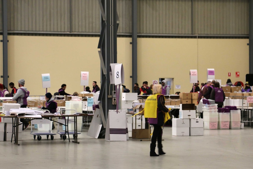 A wide shot of people sitting at tables counting votes among boxes and crates in a warehouse.