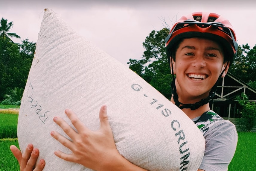 A young boy, smiling, holding a sack of fruit and wearing a bicycle helmet