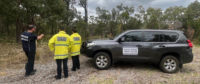 Three officers wearing clothing labelled 'police arson' look into the bush, next to car labelled 'strike force vulcan'