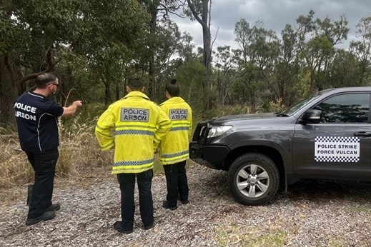 Three officers wearing clothing labelled 'police arson' look into the bush, next to car labelled 'strike force vulcan'