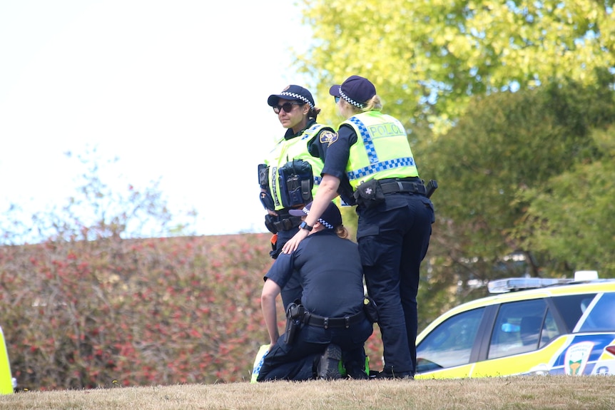 Three police officers console each other with emergency service vehicles in the background.