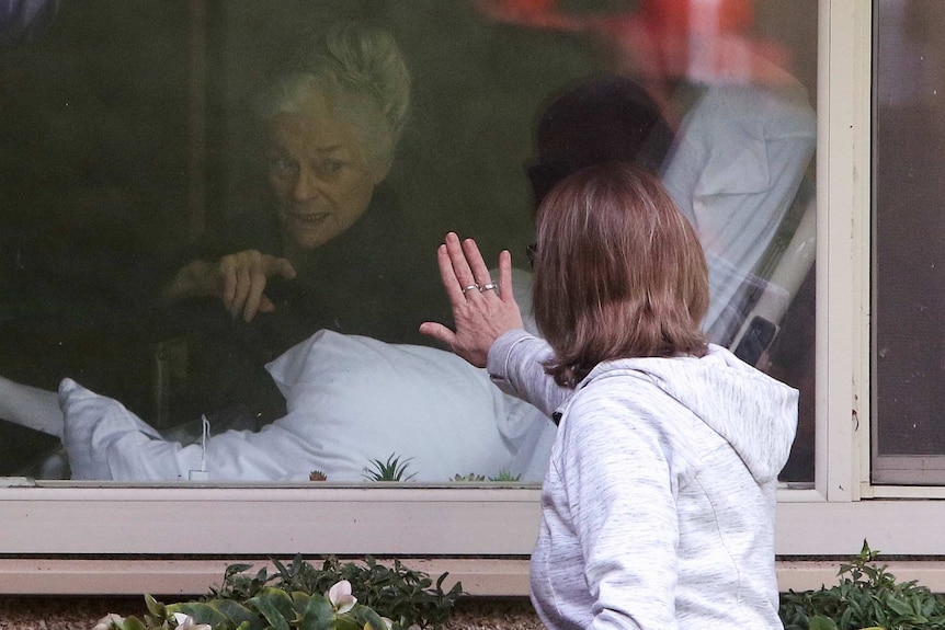 A woman with her hand pressed to a window with an older woman looking from inside