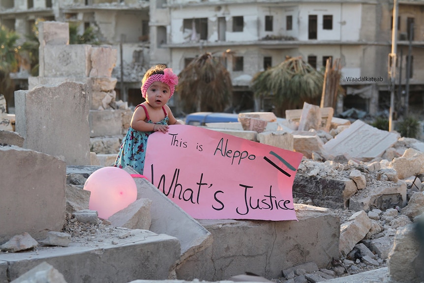A baby girl wears pink headband and stands in ruins of building and holds pink sign which reads: this is Aleppo, what's justice.