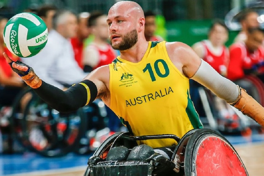 A bald man with beard playing wheelchair rugby in a yellow and green jersey holding a green and white ball with one hand