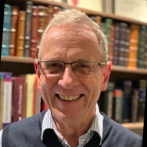 A man in glasses smiles, standing in front of a bookshelf.