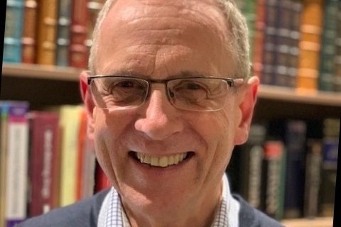 A man with glasses smiles, standing in front of a shelf.