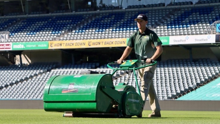 A man pushes a large green mower across sport stadium turf.