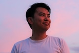 A young man stands against a pink sunset sky. 