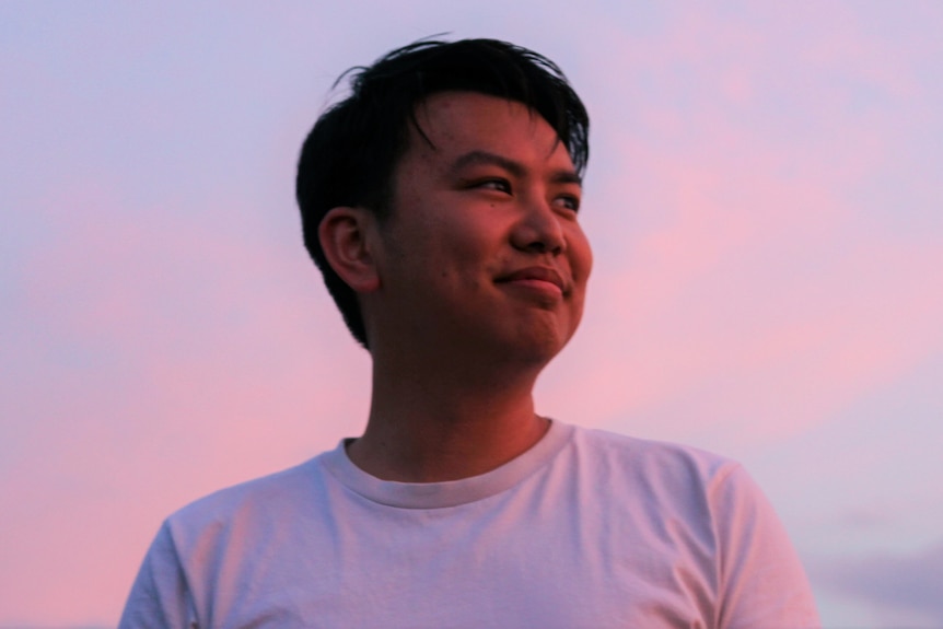 A young man stands against a pink sunset sky. 