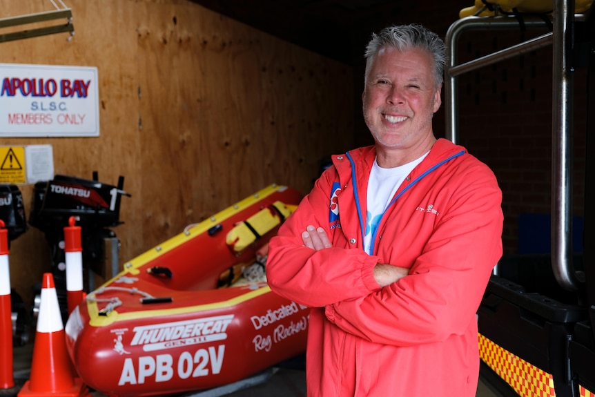 Mr Gorrie stands in a garage surrounded by life saving equipment in a red jacket smiling. 