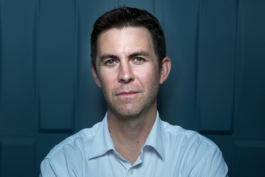 A man with brown hair, white skin and a pale blue shirt stands with arms crossed looking at the camera. 