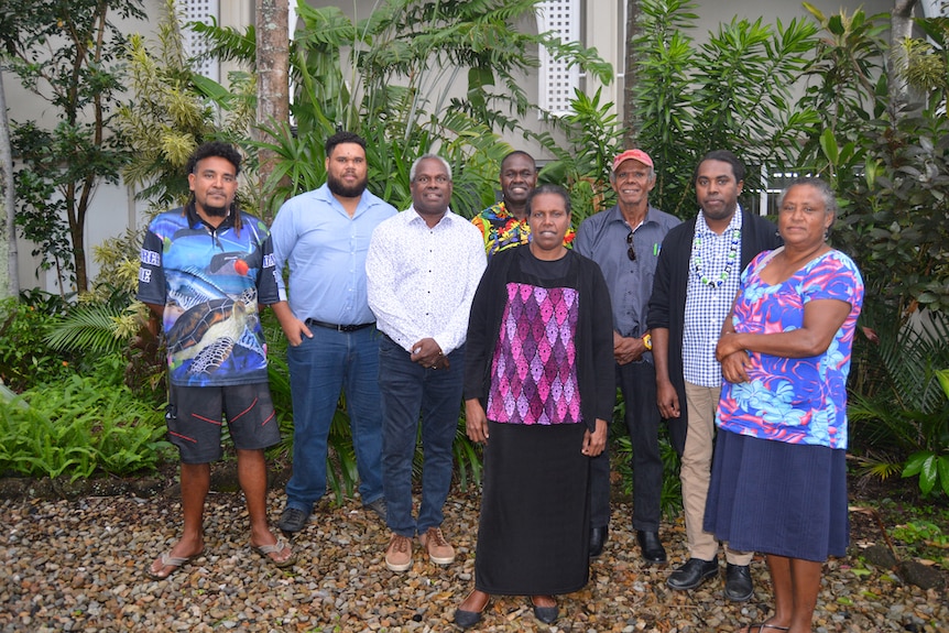 Eight Torres Strait Islander people pose proudly for the camera in front of a tropical-looking garden.